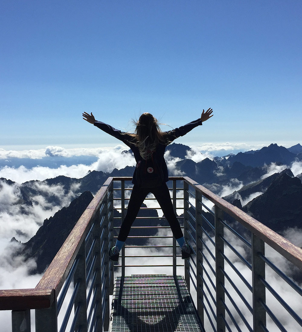 Frau hüpft auf einem Aussichtspunkt vor einem Bergpanorama in die Luft