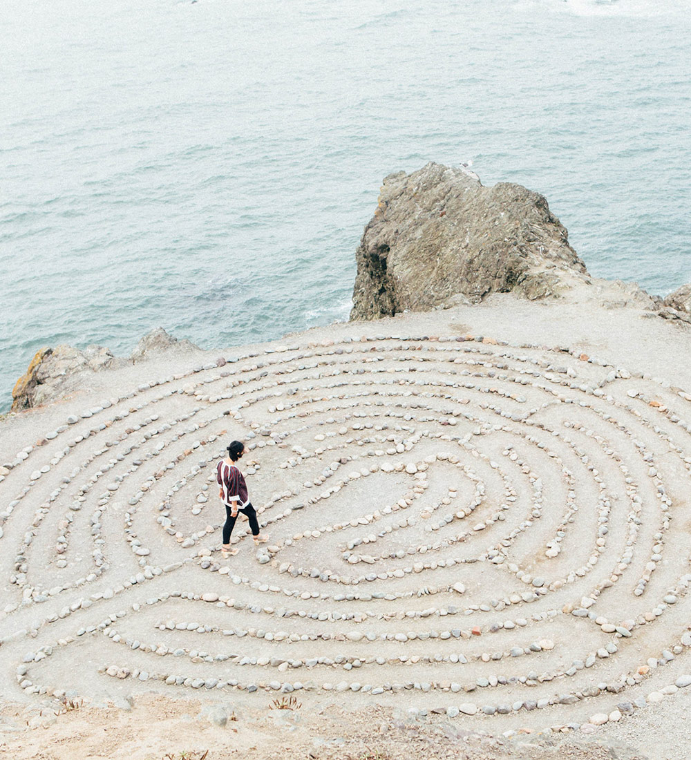 Frau läuft am Meer entlang eines aus Steinen gelegten Labyrinths